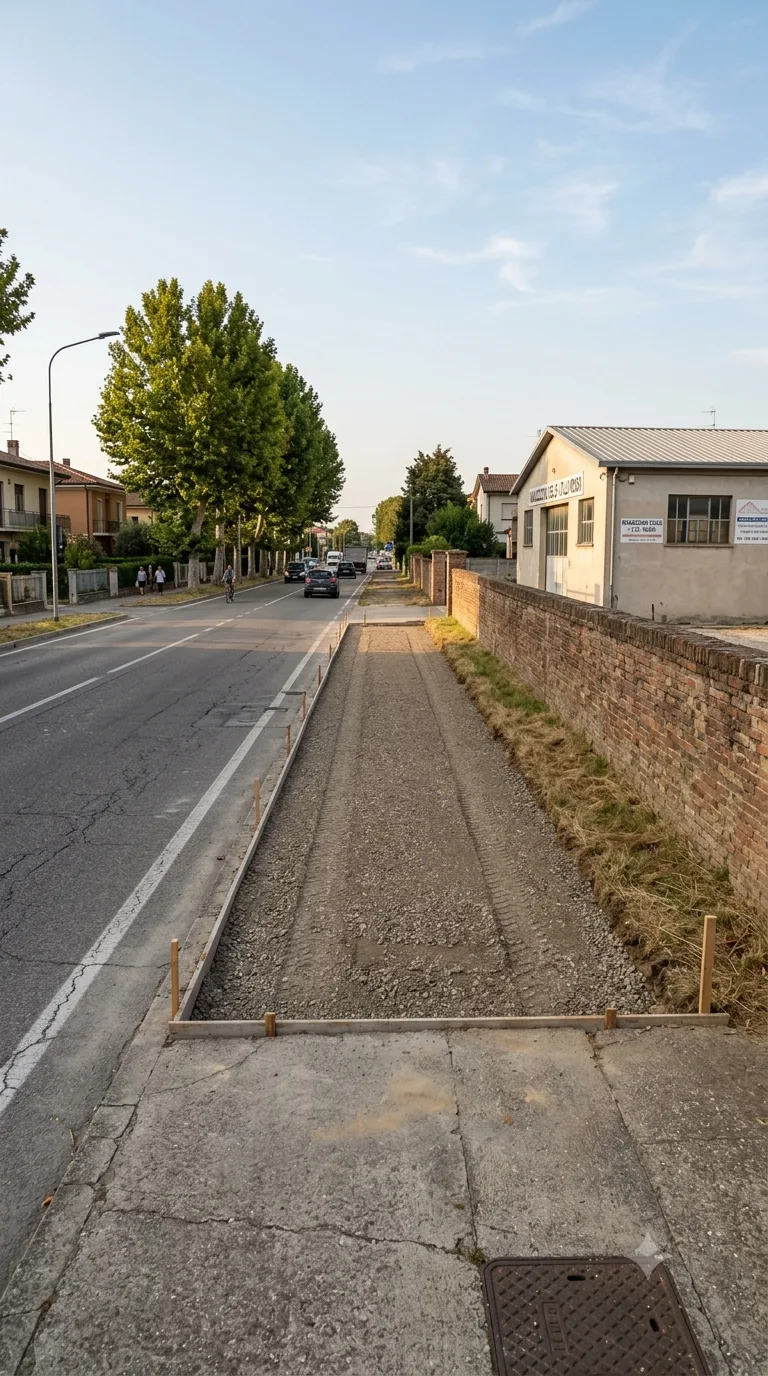 Immagine statica verticale 9:16 per YouTube Shorts, destinata a Flow Veo e a un timelapse di costruzione. Inquadratura fissa leggermente rialzata a tre quarti dal marciapiede, a circa 18 metri dal centro della fascia di servizio. A sinistra corre una strada provinciale asfaltata a due corsie con segnaletica consunta e un marciapiede in calcestruzzo; sullo sfondo del marciapiede opposto un filare di platani. A destra un muro di confine basso in laterizio di un magazzino intonacato chiaro. Al centro una fascia di servizio in ghiaia compatta, livellata, continua e libera, con erba bassa ai margini verso il muro. Pomeriggio nitido, luce laterale da sinistra, cielo azzurro pallido con nubi sottili, color grading naturale leggermente caldo. Nessun movimento di camera.