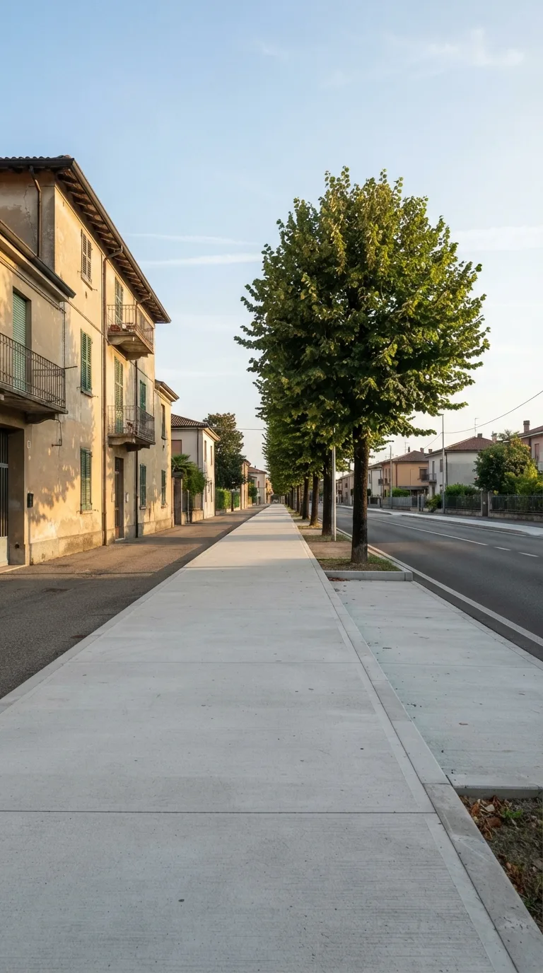 Vertical 9:16 YouTube Shorts image for Flow Veo construction timelapse. Static camera, fixed perspective, no camera movement. On a quiet urban residential street in the Italian plains, a wide concrete sidewalk runs parallel to a two-lane asphalt road. To the left stands a three-story residential building with light ochre plaster walls and simple rectangular windows. A row of mature lime trees lines the far side of the sidewalk, casting soft dappled shade. The sidewalk surface is clean concrete with a low curb; beyond the curb the asphalt road is empty. Background shows distant low houses under a clear afternoon sky with thin high clouds. Soft natural daylight from the left, slightly warm color grading, realistic photographic look. The sidewalk area in the foreground and mid-ground remains completely open and empty, showing only the existing pavement, curb, building wall and tree trunks. No construction elements present.