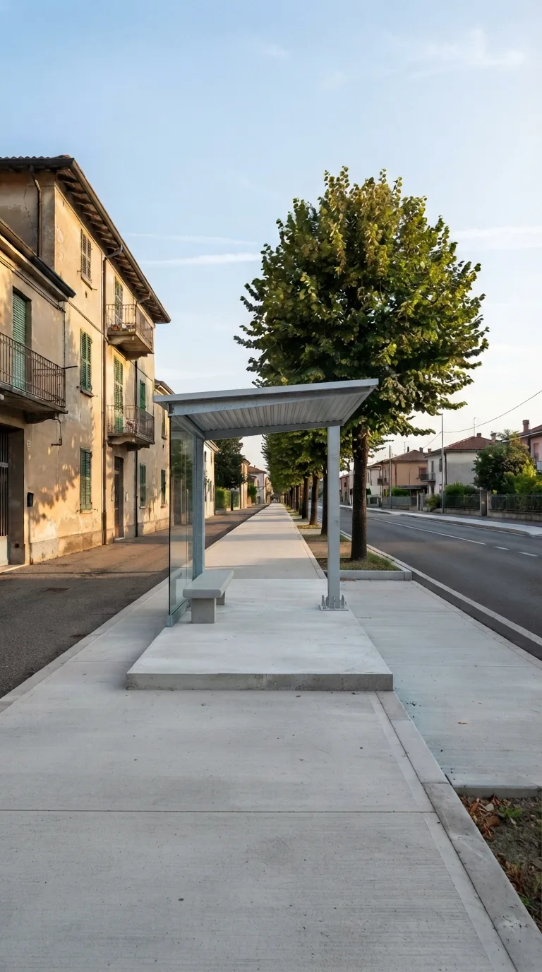 Vertical 9:16 YouTube Shorts image for Flow Veo construction timelapse. Static camera, fixed perspective, no camera movement. On a quiet urban residential street in the Italian plains, a wide concrete sidewalk runs parallel to a two-lane asphalt road. To the left stands a three-story residential building with light ochre plaster walls and simple rectangular windows. A row of mature lime trees lines the far side of the sidewalk, casting soft dappled shade. The sidewalk surface is clean concrete with a low curb; beyond the curb the asphalt road is empty. Background shows distant low houses under a clear afternoon sky with thin high clouds. Soft natural daylight from the left, slightly warm color grading, realistic photographic look. Fully finished bus shelter occupies the sidewalk: a 6 m long, 2.2 m deep, 3 m high cantilevered canopy of light-grey corrugated metal sloping toward the road, supported by two rear galvanized steel columns. A 4 m long grey concrete bench sits under the canopy. A tempered glass side panel closes the left end. The concrete base plate is flush with the sidewalk. The structure is clean, complete and ready for use, with free space around it on the sidewalk.