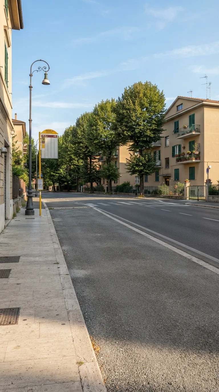 Immagine statica per Flow Veo, YouTube Shorts verticale 9:16, da usare per un timelapse di costruzione. Slargo asfaltato di un quartiere residenziale di pianura italiano, visto in prospettiva obliqua statica a livello d’uomo da circa 18 metri, inquadratura verticale centrata sulla fascia di attesa tra il marciapiede in lastre di pietra chiara in primo piano a sinistra e la corsia stradale a due sensi a destra. Sul fondo tre palazzine basse intonacate color sabbia con persiane verdi; un filare di tigli adulti allinea il lato opposto della strada; un lampione esistente in ghisa scura resta sul bordo del marciapiede; una palina segnaletica gialla già piantata marca la fermata. L’asfalto dello slargo è liscio, grigio scuro, con una fascia di ghiaia fine verso il filare. Luce pomeridiana laterale da sinistra, cielo azzurro con nubi sottili, color grading naturale leggermente caldo. Nessun movimento di camera.