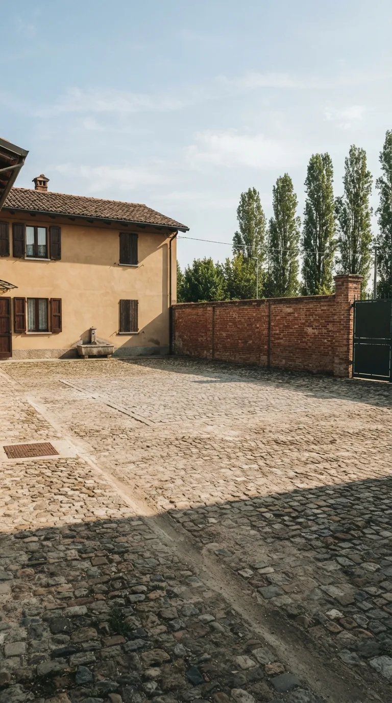 Static vertical 9:16 YouTube Shorts still for a construction timelapse. Eye-level camera about 1.6 meters high, three-quarter view from the courtyard, standing about 18 meters from the center of the open cobbled yard, looking toward the rear of the lot. Horizon sits mid-frame. Foreground: worn cobblestone pavement of mixed grey and brown stones with irregular joints and a faint tire-polished track running from the lower left toward the center. Center: a clear rectangular patch of the same cobblestones, slightly flatter, with no excavation and no piled materials. Left background: a two-story plastered house in warm ochre with dark timber shutters and a terracotta tiled roof. Right background: a continuous red-brick boundary wall about 2.2 meters high with weathered mortar. Far background: a row of poplars beyond the wall and a pale blue late-morning sky with thin white clouds. Existing elements stay fixed: a dark-green steel pedestrian gate in the brick wall at the far right, a cast-iron drain cover near the house corner on the left, and a low stone water trough against the house wall. Natural late-morning sunlight from the left, crisp contact shadows, photographic color grading, slightly warm, enough depth of field to keep house, yard and wall sharp. No people, no vehicles, no construction gear.
