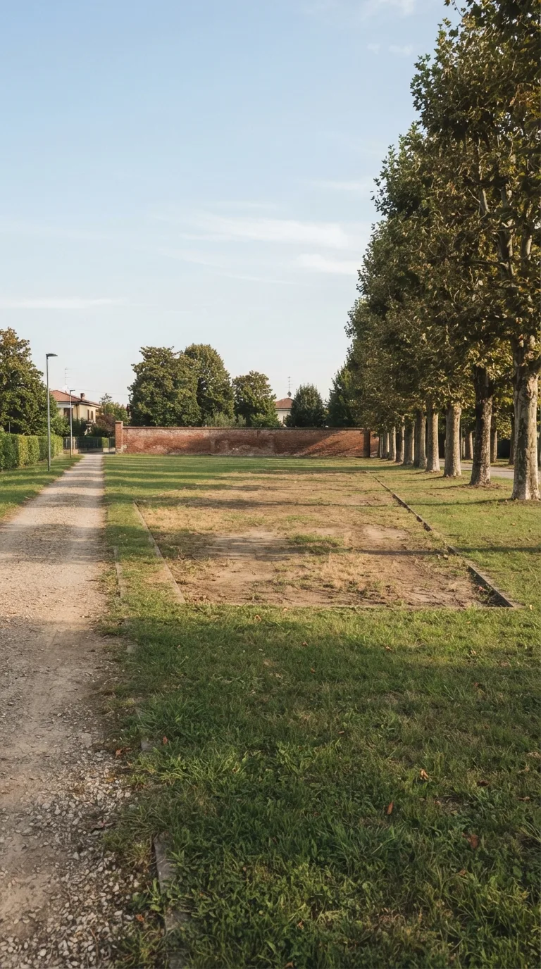 Still photograph for a YouTube Shorts vertical 9:16 construction timelapse. Neighborhood park on the Po plain. A packed gravel path runs along the left side of the frame; a row of mature plane trees stands on the right; a low existing brick boundary wall closes the far background; unused lawn occupies the near foreground and the margins outside a central worn rectangle of packed earth and thin grass about sixteen metres long and ten metres wide. Static camera about 2.2 metres high, three-quarter view from the near-left corner looking toward the far-right trees, horizon in the upper third, generous margin around the central ground. Clear afternoon light from the left, pale blue sky with thin clouds, natural slightly warm color grading, photographic depth of field that keeps path, trees and wall sharp. The central ground is only packed earth with sparse dry grass, no paving, no excavation, no materials stacked. No text, logos or watermarks.