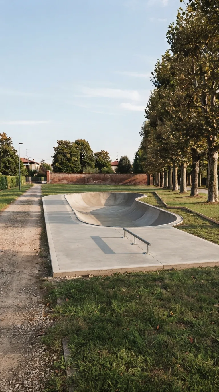 Still photograph for a YouTube Shorts vertical 9:16 construction timelapse. Neighborhood park on the Po plain. A packed gravel path runs along the left side of the frame; a row of mature plane trees stands on the right; a low existing brick boundary wall closes the far background; unused lawn occupies the near foreground and the margins around the finished work. Static camera about 2.2 metres high, three-quarter view from the near-left corner looking toward the far-right trees, horizon in the upper third, generous margin around the structure. Clear afternoon light from the left, pale blue sky with thin clouds, natural slightly warm color grading, photographic depth of field that keeps path, trees and wall sharp. A compact skatepark occupies the centre: a smooth grey concrete kidney bowl 8 metres wide, 6 metres long and 1.4 metres deep with continuous zinc-coated steel pipe coping on the rim, joined toward the camera to a flat grey concrete pad 4 metres deep and 8 metres wide. A 3-metre steel grind rail sits on low mounts along the centre of the pad. A low concrete curb frames the 12 by 8 metre lot. The site is clean, empty of workers and plant, ready for use. No text, logos or watermarks.