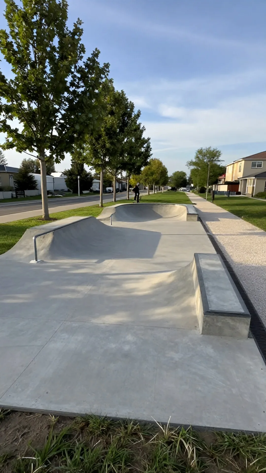 Immagine statica per YouTube Shorts verticale 9:16, da usare in un timelapse di costruzione. Parco di quartiere di pianura in una giornata pomeridiana limpida. Camera fissa a circa 2 metri dal suolo, prospettiva obliqua a livello d’uomo da circa 16 metri dallo slargo centrale, orizzonte a un terzo dall’alto. In primo piano erba bassa e un bordo di terra battuta; al centro la pista finita con spazio libero attorno; a sinistra un filare di platani adulti con foglie verdi e tronchi grigi; a destra un sentiero in ghiaia chiara che corre verso il fondo; sullo sfondo palazzine residenziali basse intonacate e un tratto di cielo azzurro con nubi sottili. Luce naturale da sinistra-alto, ombre di contatto nette e corte, color grading fotografico naturale leggermente caldo. La platea in calcestruzzo grigio liscio misura 12 metri per 8 metri; sul lato corto lontano il quarter pipe in calcestruzzo alto 1,2 metri e largo 4 metri ha transizione continua e copings in tubo d’acciaio; sul lato sinistro un rail in acciaio zincato lungo 4 metri poggia su due basette in calcestruzzo; sul lato destro un ledge in calcestruzzo alto 40 centimetri e lungo 5 metri affianca il piano; una griglia di drenaggio corre lungo il lato destro. Un solo skater con casco e skateboard percorre il rail al centro-sinistra, in scala realistica, senza coprire il quarter pipe. Cantiere assente, superfici pulite.