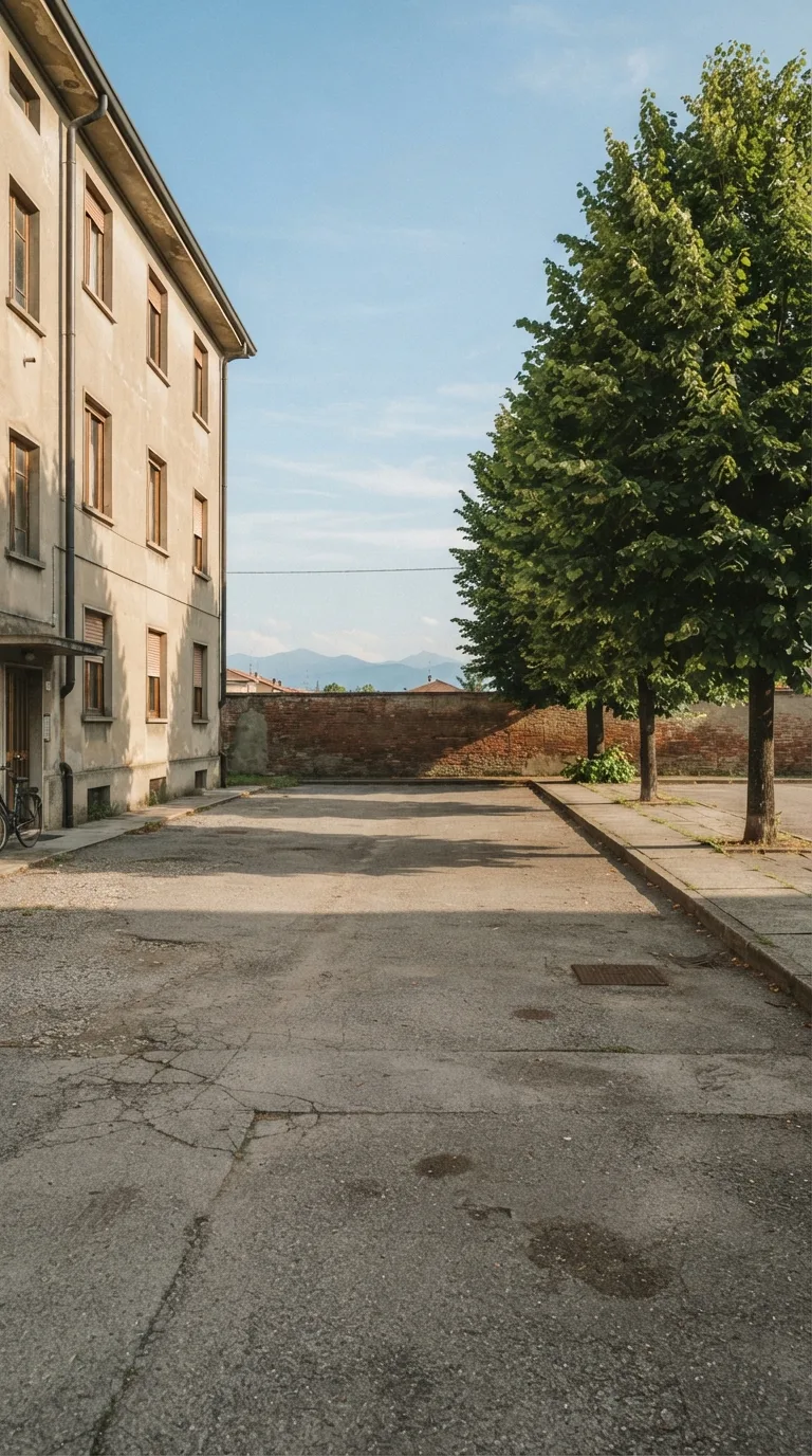Immagine statica per YouTube Shorts verticale 9:16, destinata a un timelapse di costruzione. Cortile asfaltato di un isolato residenziale di pianura italiana. Inquadratura verticale fissa, camera a circa 1,6 metri da terra, prospettiva obliqua da sud-ovest a circa 14 metri dal centro dello slargo. L’orizzonte taglia il terzo superiore. In primo piano a sinistra e in basso l’asfalto grigio consunto con giunti e macchie chiare; al centro uno slargo asfaltato libero, livellato, senza scavi. A sinistra, in secondo piano, la facciata laterale di un edificio residenziale a tre piani intonacato beige con quattro finestre allineate e un pluviale scuro. A destra un filare di tre tigli adulti con foglie verdi dense e un marciapiede in lastre di cemento. Sul fondo un muro di recinzione basso in laterizio e il cielo. Luce pomeridiana chiara da sinistra, ombre di contatto nette e corte, cielo azzurro con nubi sottili, color grading fotografico naturale leggermente caldo, profondità di campo sufficiente a leggere asfalto, edificio e tigli.